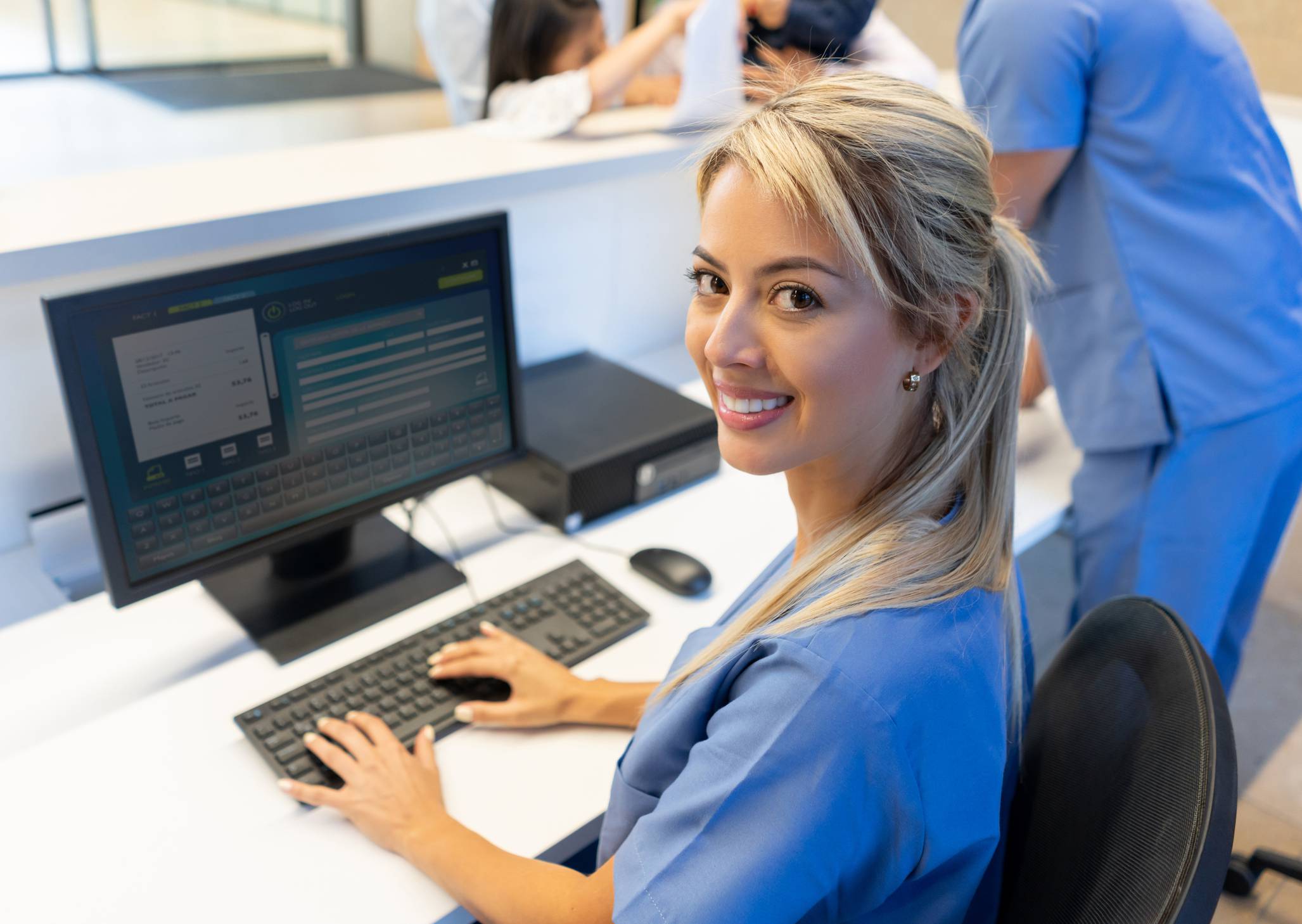 Na foto, uma moça com um jaleco azul aparece usando um computador.